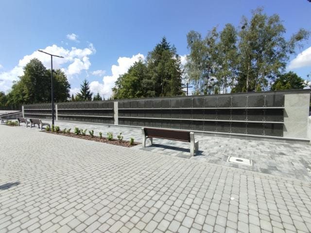 Obrazek przedstawia utwardzoną ścieżkę z&nbsp;ławkami i&nbsp;latarniami po bokach. Wzdłuż jednej strony ciągnie się długa ściana z&nbsp;wieloma rzędami tabliczek lub&nbsp;wnęk, co może sugerować, że&nbsp;jest to część memoriału lub&nbsp;kolumbarium. Teren jest zadbany, z&nbsp;niewielkimi roślinami oraz&nbsp;drzewami w&nbsp;tle. Niebo jest częściowo zachmurzone, ale&nbsp;przeważnie słoneczne, co wskazuje na&nbsp;pogodny dzień.